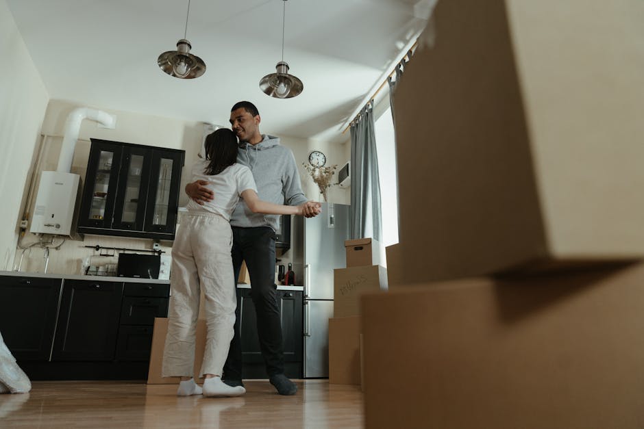 A man and a woman are inside a room, embracing with smiles during a home relocation. The woman, dressed in light-colored trousers and a white top, is turned towards the man, who is dressed in dark trousers and a grey hoodie. They stand next to a black kitchen unit with glass-fronted cabinets, with boxes and packing materials such as cardboard boxes and plastic-wrapped items visible nearby on the wooden floor. The room has a light, neutral wall color with a window draped with light curtain panels, allowing natural light to fill the space. Overhead, there are three modern ceiling light fixtures. The scene captures a moment during the loading process of furniture and packed belongings, with several moving boxes stacked near the window, indicating an ongoing house move. The setting includes elements typical of a residential property, and the overall image reflects the packing and moving activities associated with house removals, as provided by Croydon Movers.