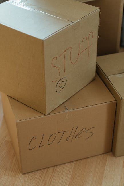 A close-up image showing three cardboard moving boxes inside a property, likely on the floor near a wall or doorway. The top box, marked with red marker as 'STUFF' and a small smiley face, is partially visible. The middle box, labeled 'CLOTHES' in black marker, is positioned underneath the first. The boxes appear to be sealed with packing tape and are made of brown kraft cardboard, used for packing belongings during household moves. The floor surface is wooden, and natural light illuminates the scene, suggesting daytime. The image captures part of a home relocation process, illustrating organized packing and preparation for furniture transport and moving logistics, with boxes ready for loading onto a van or storage area. Croydon Movers, a local removals company, may be involved in the packing and loading process depicted.