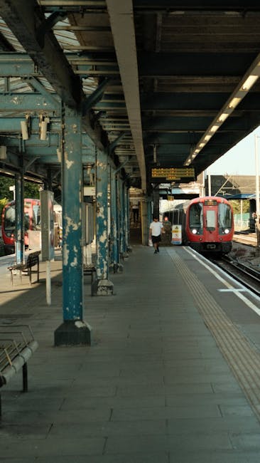 The image shows a train station platform with a covered, weathered metal canopy supported by blue-painted steel columns. Under the canopy, there are several benches, some of which are empty. A person dressed in light clothing is walking along the platform toward a red train that is stopped at the station. The train has large windows and is positioned alongside the platform, with another carriage visible further down the tracks. The platform surface is made of concrete, featuring tactile paving strips near the train doors for accessibility. The environment is lit with natural daylight, with additional lighting fixtures mounted beneath the canopy. The scene captures a quiet moment during a typical weekday, illustrating a public transport setting suitable for house removals or relocation services, where careful planning for transport, loading, and passenger movement is essential. Croydon Movers' services often support clients in logistical planning for home relocation, including transport on train networks such as this one, with considerations for packing and loading between train journeys and home move planning.