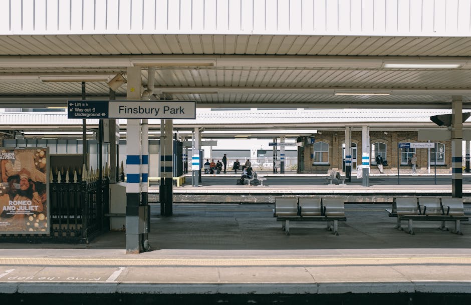 A view of Finsbury Park railway station platform with a covered waiting area, showing multiple metal benches arranged along the edge of the platform. The platform features a white and black striped signage pole with a sign indicating 'Finsbury Park,' and additional signs directing to the lift and underground exits. In the background, there are several passengers sitting on benches or walking along the platform, some with luggage. To the left, a poster for 'Romeo and Juliet' is visible on a nearby wall. The scene is captured during daylight hours, with natural light illuminating the station environment. This setting illustrates typical elements of railway station platforms, relevant to home relocation and furniture transport in the context of local moving or packing services offered by Croydon Movers, with emphasis on the transportation logistics involved in moving within or from these transit locations.