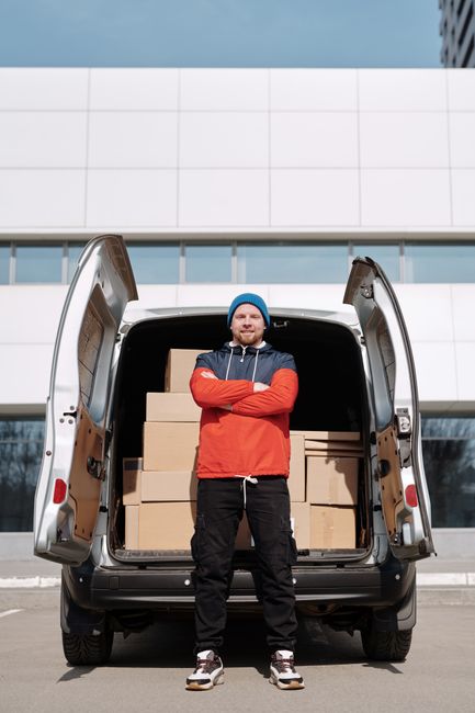 A man wearing a blue beanie, red and navy jacket, and black pants stands with arms crossed in front of an open van loaded with multiple cardboard boxes for a home relocation. The van is parked outside