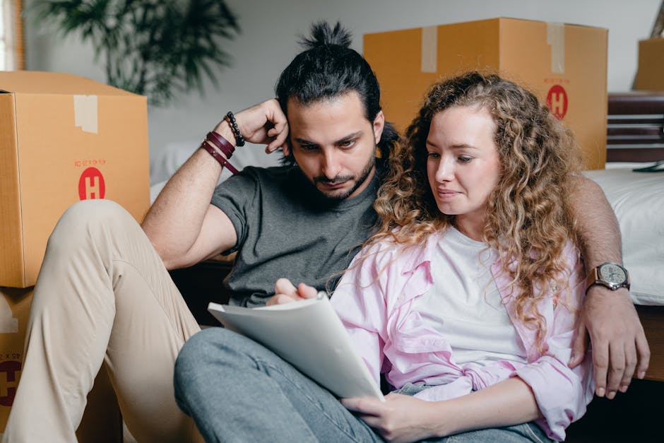 A young man and woman sit on the floor inside a property surrounded by packed moving boxes made of cardboard, ready for a home relocation. The man, with dark hair tied back and wearing a grey t-shirt and beige trousers, is leaning against a cardboard box with his right arm, while the woman, with curly light brown hair and wearing a pink shirt and grey jeans, is holding a clipboard or document and looking at it. They are positioned near a doorway that leads to the outside, where additional boxes and a vehicle for furniture transport may be present, indicating an active loading process. The room features typical home furnishings, including a bed, and the scene is well-lit with natural light. [COMPANY_NAME], as part of the house removals service, is involved in coordinating packing and moving logistics, ensuring the careful handling of furniture and boxes during the moving process.